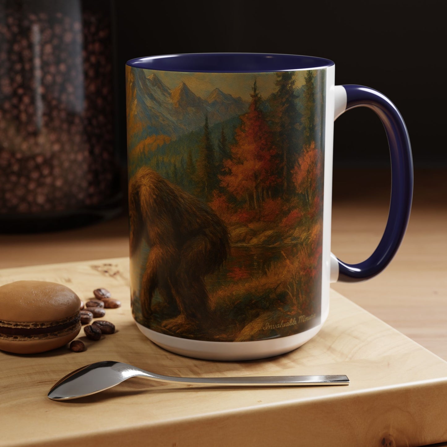 15oz Bigfoot River Scene ceramic coffee mug with navy blue handle and interior — right-side view showing autumn forest, mountain, and river reflections. Styled on a wooden table with coffee beans, macaron, and spoon for cozy lifestyle presentation. Boutique drinkware by Invaluable Minutia. InvaluableMinutia.com